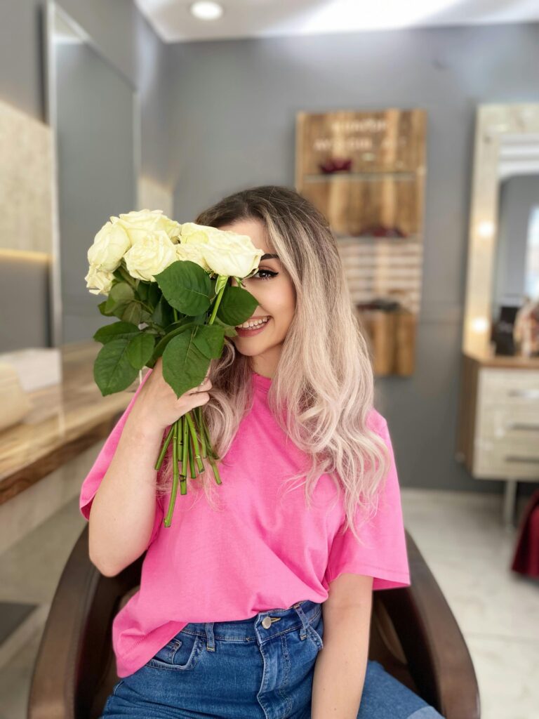 A happy woman in a pink shirt holds white roses, smiling indoors. Bright and cheerful ambiance.